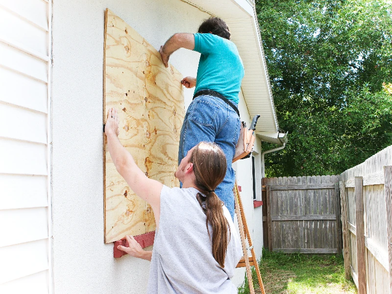 Two people boarding up a window on a house with a large plywood sheet. emergency board up service